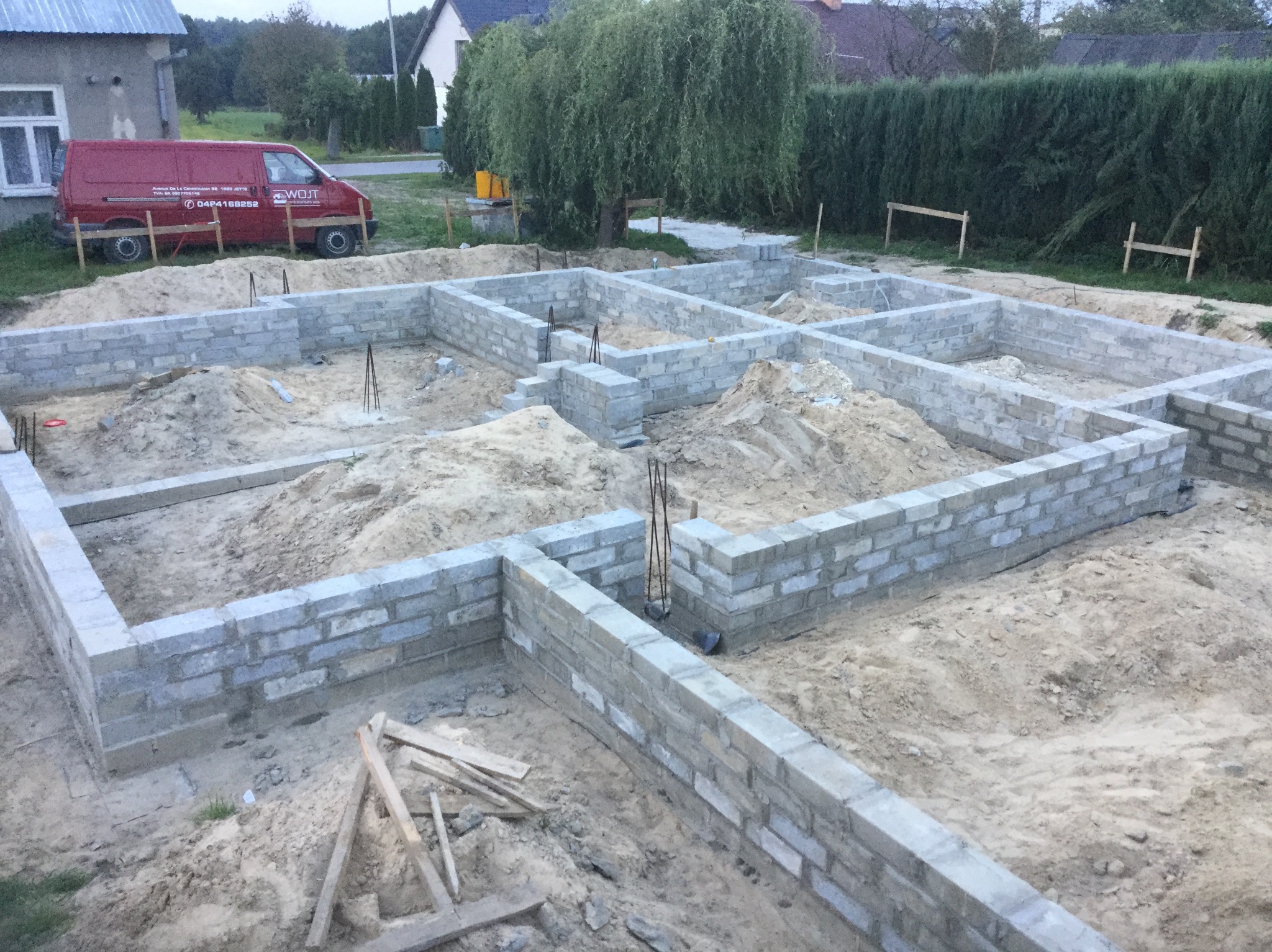 Fundamenty domu z bloczków betonowych, widok z góry. W tle zaparkowany czerwony bus firmy budowlanej i zabudowa jednorodzinna.
