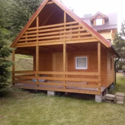 Drewniany domek letniskowy z zadaszonym tarasem, widoczny z zewnątrz, na betonowych fundamentach, z czerwoną blachodachówką, otoczony zielenią.