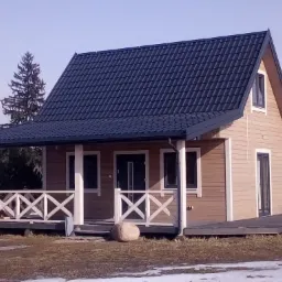 Gotowy, parterowy budynek z drewnianą elewacją i ciemnym dachem z blachodachówki, widoczny z przodu, z charakterystycznym balustradowym wykończeniem werandy i dużym kamieniem przed nią.