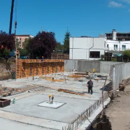 Wykop pod fundamenty nowego budynku z widocznym zbrojeniem, szalunkami i pracownikami budowlanymi w tle, z dźwigami i budynkami w oddali.