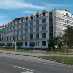 Wielopiętrowy budynek w trakcie ocieplania elewacji, widoczne rusztowania i siatka ochronna, częściowo zamontowana izolacja termiczna, okna wstawione, tło: błękitne niebo i drzewa.