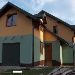 Dwukondygnacyjny dom z garażem, elewacja w kolorach zieleni i beżu, widoczne haki na ścianie, stos desek przed budynkiem.