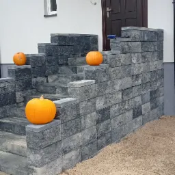 Schody wejściowe do domu wyłożone szarą kostką brukową, udekorowane trzema pomarańczowymi dyniami, z widocznym fragmentem chodnika z kostki brukowej i piaskiem.