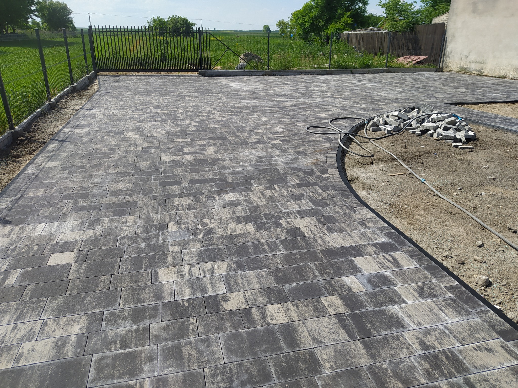 Ułożona kostka brukowa w odcieniach szarości, z widoczną stertą kostek do ułożenia i ogrodzeniem w tle.