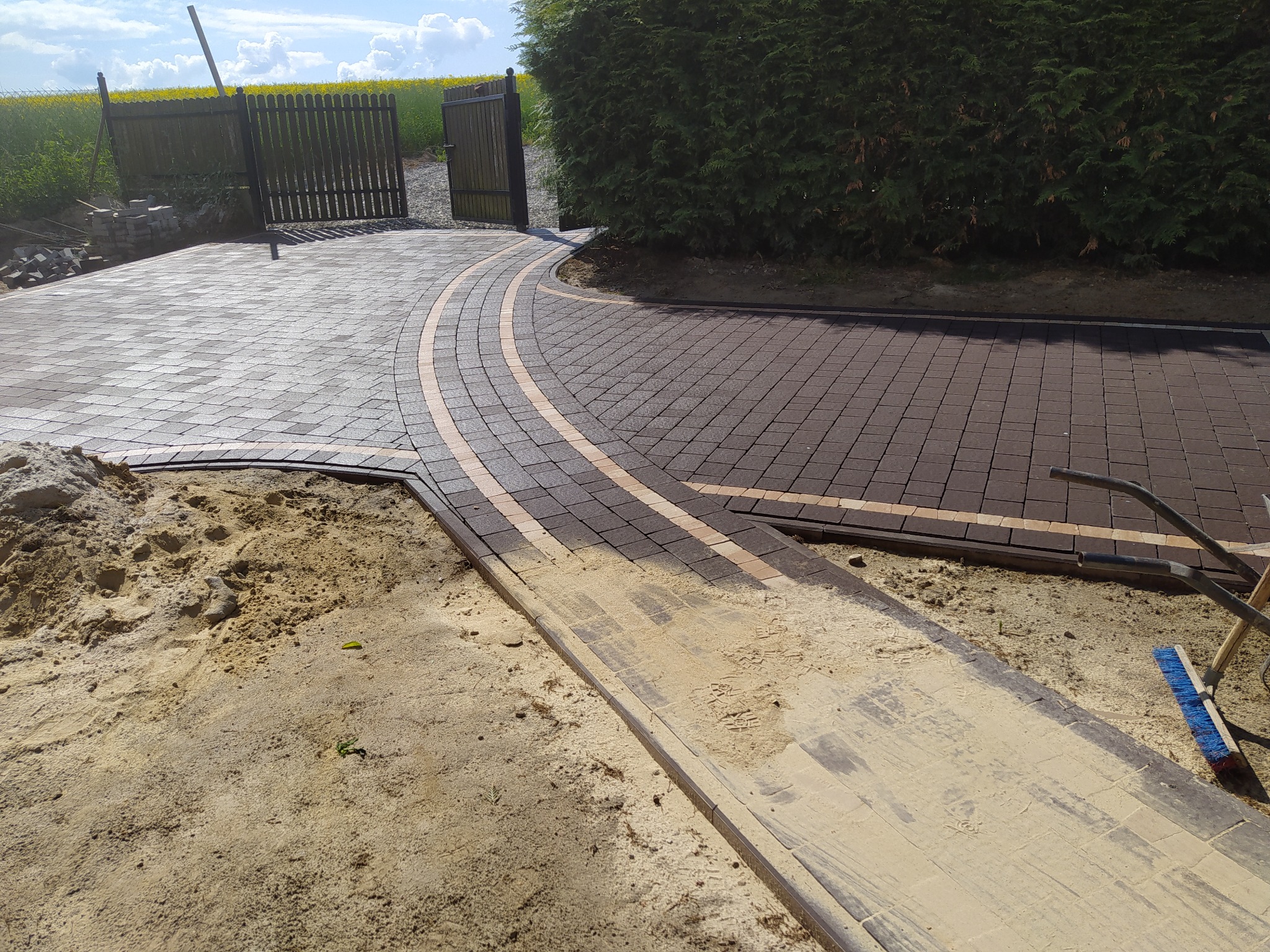 Nowo ułożony, zakrzywiony podjazd z kostki brukowej w odcieniach szarości i brązu, z widocznym piaskiem do wypełnienia spoin i szczotką do zamiatania, prowadzący do otwartej bramy w tle z kwitnącym...