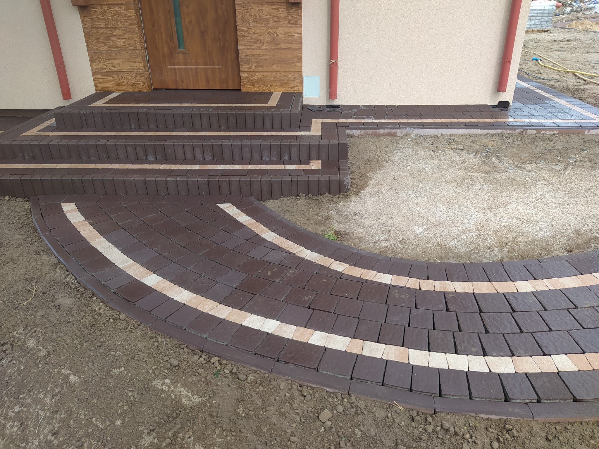 Brązowa kostka brukowa ułożona w łukowaty wzór i schody wejściowe do domu, z beżowymi akcentami, na tle beżowej elewacji budynku i widocznym fragmentem trawnika.