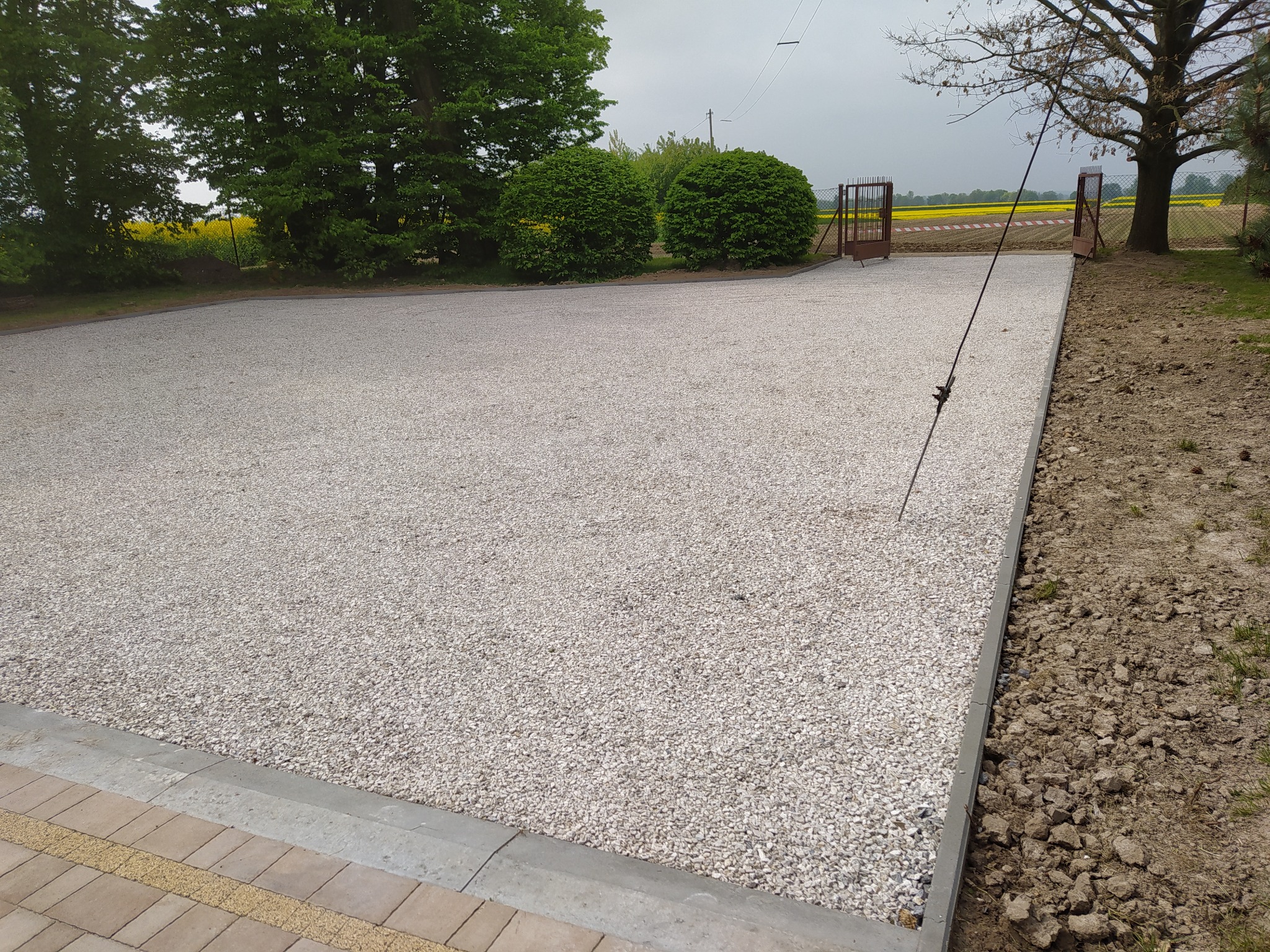 Ukończona powierzchnia z białego grysu otoczona betonową obwódką, widok na brązową bramę wjazdową i pole rzepaku w tle.