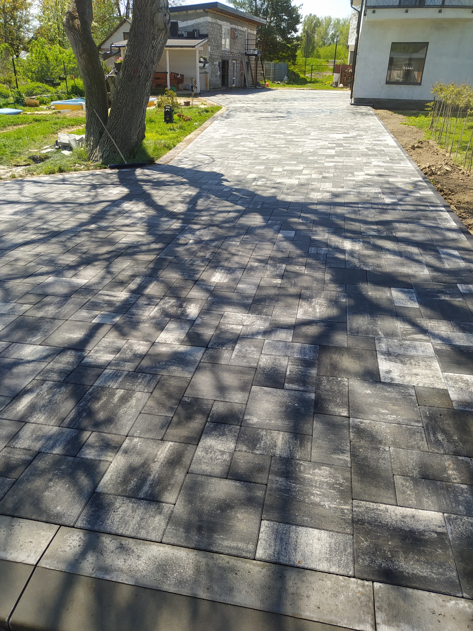 Ułożona, szara kostka brukowa na podjeździe, częściowo zacieniona przez drzewo, widok na dom w tle.