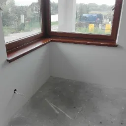 Wnętrze pokoju po malowaniu na biało, z drewnianym parapetem i szarym betonowym podłożem, widoczne przewody wystające ze ściany.