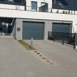Nowoczesny dom bliźniak z szarą kostką brukową, grafitowymi bramami garażowymi i ogrodzeniem panelowym; widoczna skrzynka gazowa w żółtym kolorze.