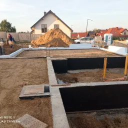 Przygotowany fundament pod budynek jednorodzinny, z izolacją pionową i poziomą, widoczne rury kanalizacyjne, w tle sterta piasku i budowa.