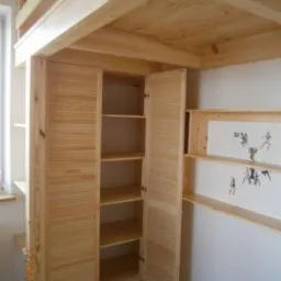 Drewniana szafa wnękowa z lamelowymi drzwiami, zintegrowana z konstrukcją łóżka na antresoli, z otwartymi półkami po prawej stronie, wykonana z jasnego drewna.