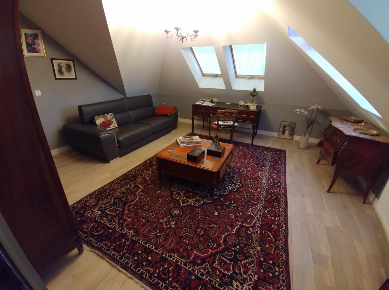 Pokój na poddaszu z szarą ścianą, skórzaną sofą, biurkiem pod oknami dachowymi i orientalnym dywanem w odcieniach czerwieni i granatu.