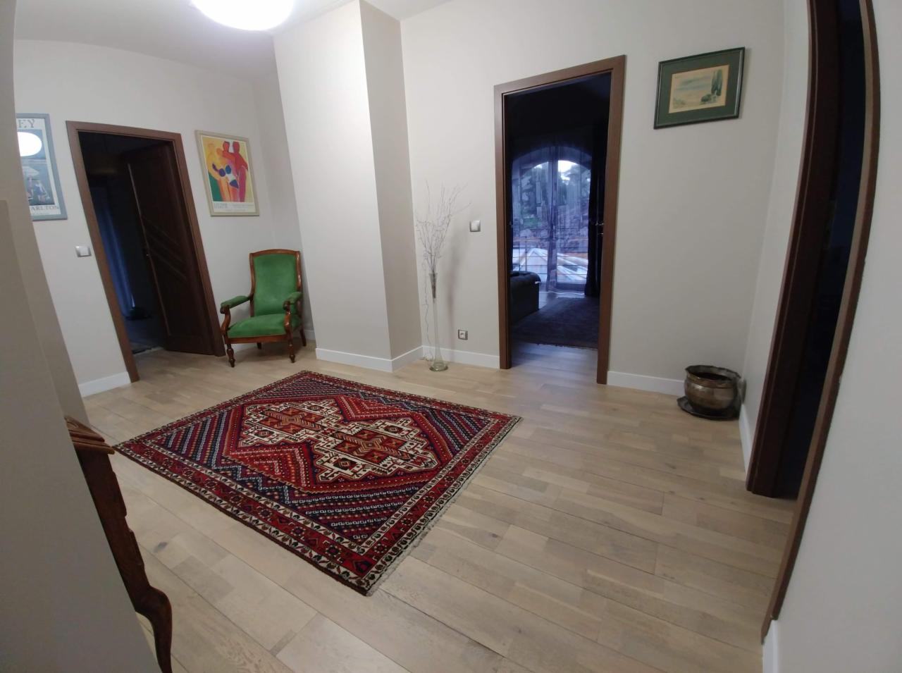 Jasny przedpokój z parkietem, orientalnym dywanem, zielonym krzesłem i dwoma obrazami na ścianach. W głębi widoczne drzwi z łukowym przeszkleniem.