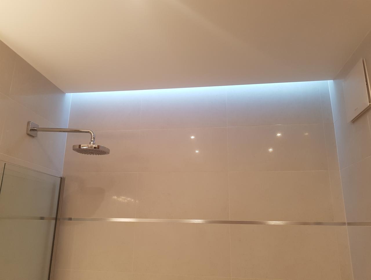 Wykończona łazienka z deszczownicą, podświetleniem LED w suficie i jasnymi, kwadratowymi płytkami ceramicznymi z delikatnym połyskiem.