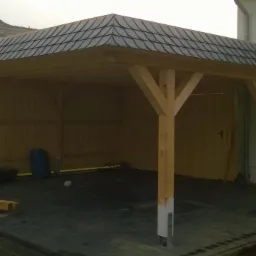 Drewniana wiata garażowa z dachem pokrytym ciemną dachówką ceramiczną, widoczne elementy konstrukcyjne z drewna oraz częściowo pomalowane na biało słupy nośne, na podłożu kostka brukowa.