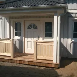 Szary dom szkieletowy z drewnianym gankiem w trakcie budowy, widoczne elementy konstrukcyjne i instalacyjne.