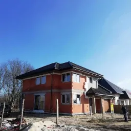 Dwupiętrowy dom w trakcie budowy z czarnym dachem, widoczne ślady prac budowlanych na posesji, piasek i ogrodzenie tymczasowe.