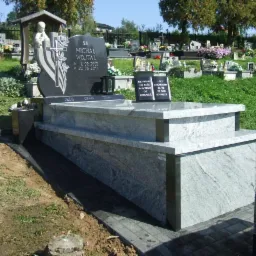 Granitowy nagrobek z rzeźbą anioła i tablicą inskrypcyjną na tle cmentarza, z nową kostką brukową wokół, w słoneczny dzień.