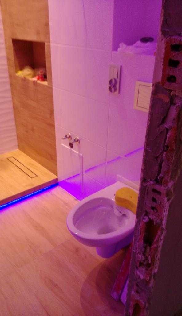 Niedokończona toaleta w łazience z podświetlanym brodzikiem i fragmentem muru w tle, oświetlona fioletowym światłem.