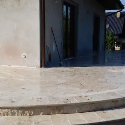 Wyłożony jasnym kamieniem taras z dwoma półokrągłymi stopniami, przylegający do budynku z dużymi oknami i widoczną miotłą opartą o ścianę.