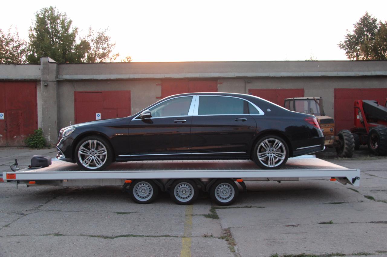 Czarna limuzyna Mercedes-Maybach 4Matic na niskopodwoziowej lawecie z trzema osiami, na tle betonowego placu z budynkiem garażowym i maszynami rolniczymi w tle.