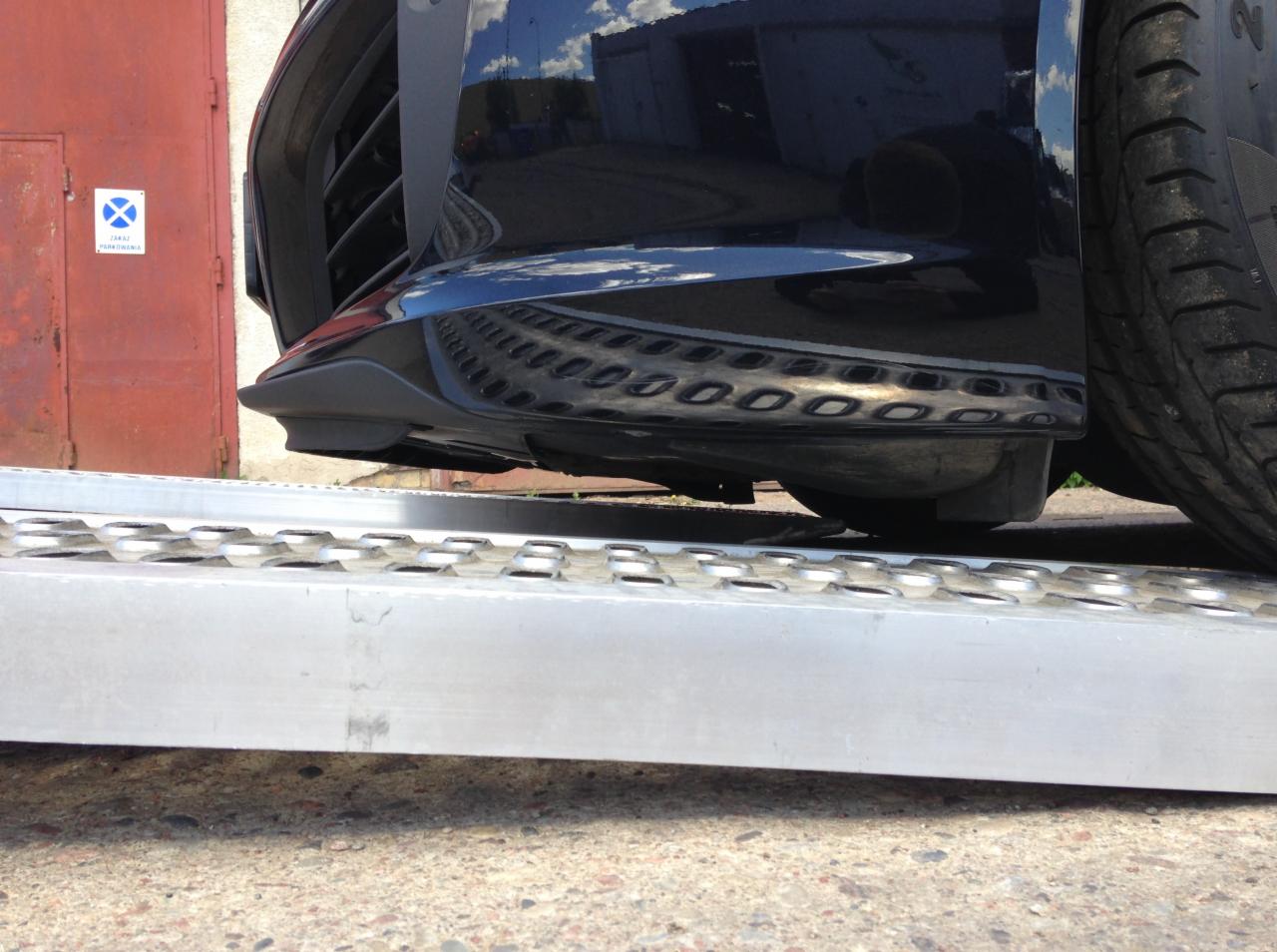 Niski kąt ujęcia czarnego samochodu na metalowej rampie, z widocznym odbiciem nieba i budynku w karoserii. Czerwone drzwi i znak zakazu parkowania w tle.