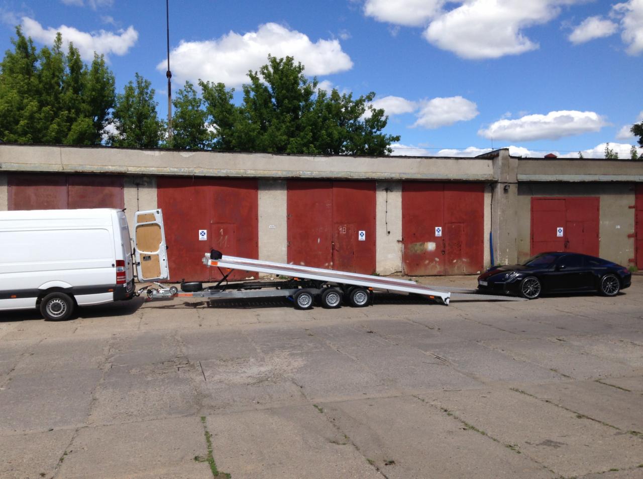 Biały van z otwartymi tylnymi drzwiami podłączony do niskopodwoziowej przyczepy, na którą wjeżdża czarny samochód sportowy; w tle rząd garaży z czerwonymi drzwiami.