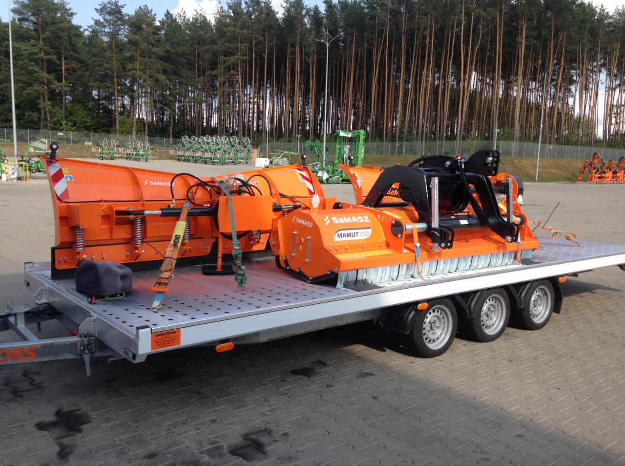 Pomarańczowe maszyny rolnicze marki Samasz, w tym pług śnieżny i kosiarka bijakowa Mamut 250, zabezpieczone pasami transportowymi na niskopodwoziowej przyczepie z trzema osiami, na tle parkingu...