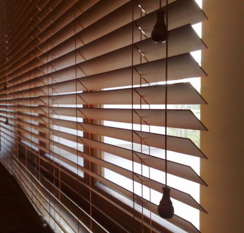 Częściowo przymknięte drewniane żaluzje poziome w oknie, widoczne sznurki z ozdobnymi uchwytami do regulacji.