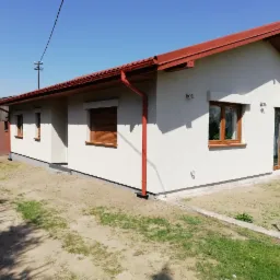 Jednopiętrowy dom z jasną elewacją i czerwoną dachówką, widoczne brązowe okna, rynny i rolety, przed domem trawnik i fragment podjazdu.