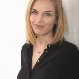 Portret uśmiechniętej blondynki w czarnej koszuli na tle białej ściany. Kobieta ma niebieskie oczy i delikatny makijaż. Widoczny naszyjnik z rubinowym kamieniem.