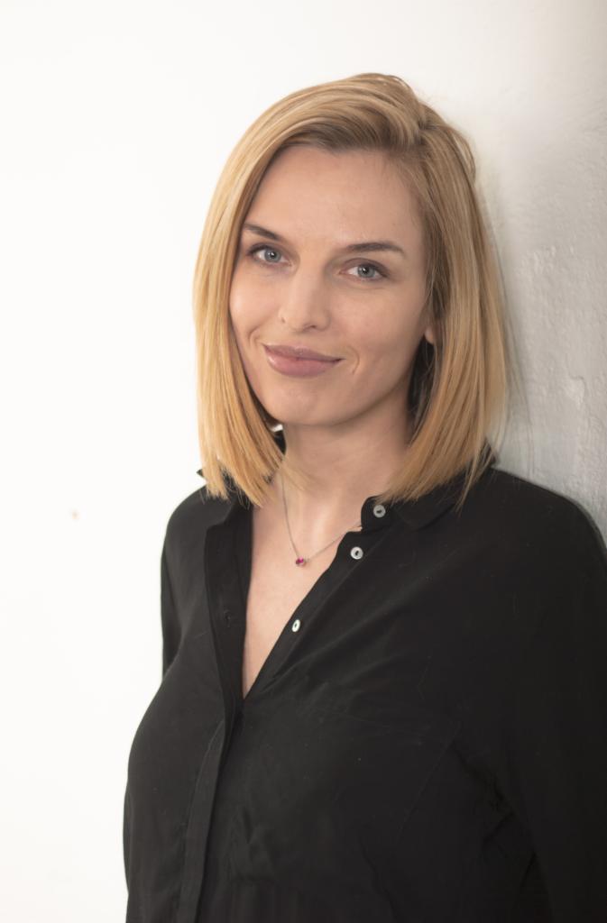 Portret uśmiechniętej blondynki w czarnej koszuli na tle białej ściany. Kobieta ma niebieskie oczy i delikatny makijaż. Widoczny naszyjnik z rubinowym kamieniem.