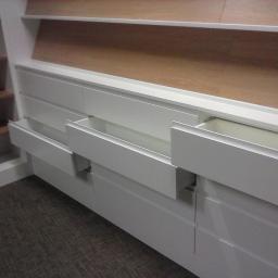 BMP Adam Prandzioch - Biała zabudowa z szufladami z częściowo wysuniętymi frontami, widoczna w Lublińcu, z drewnianą półką powyżej, na tle ciemnej podłogi.