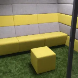 Wyciszona kabina biurowa z żółto-szarymi panelami ściennymi, modułową sofą i pufą na zielonej wykładzinie imitującej trawę, widoczna przez częściowo otwarte szklane drzwi.