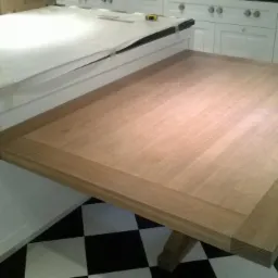 Wysuwany drewniany blat roboczy zamontowany do wyspy kuchennej z białymi frontami, widoczna czarno-biała podłoga w szachownicę.