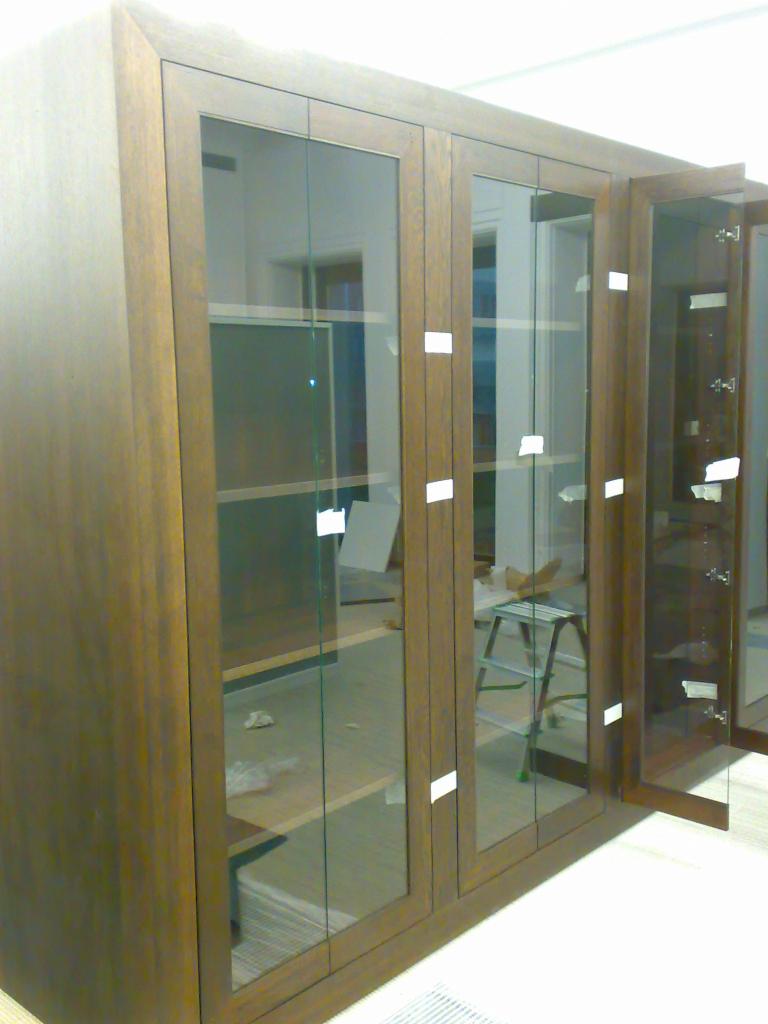 Drewniana witryna z przeszklonymi drzwiami, w trakcie montażu, widoczne zabezpieczenia na szkle i drabinka w tle.