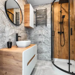 Nowoczesna łazienka z marmurowymi płytkami, drewnianą szafką pod umywalką z białą umywalką nablatową, czarną armaturą, kabiną prysznicową z drewnopodobnymi panelami oraz grzejnikiem drabinkowym.
