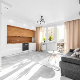 Jasne wnętrze salonu z aneksem kuchennym, białe szafki z drewnianymi frontami, szara sofa z poduszkami w gazety, szare płytki podłogowe, widok z balkonu.