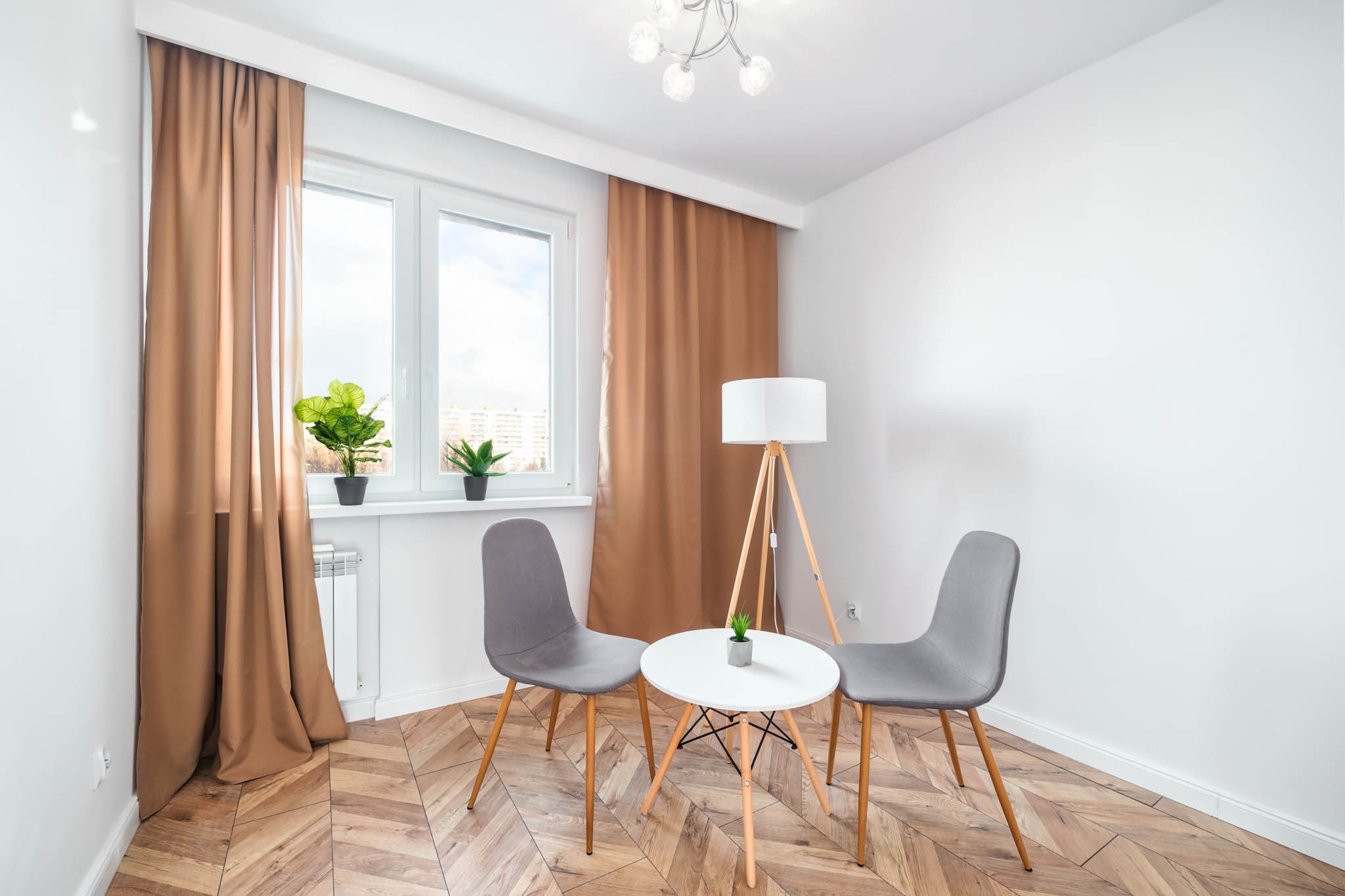 Jasne wnętrze pokoju z drewnianą podłogą ułożoną w jodełkę, brązowe zasłony przy oknie, dwie szare krzesła i okrągły biały stolik z lampką na trójnogu w tle.
