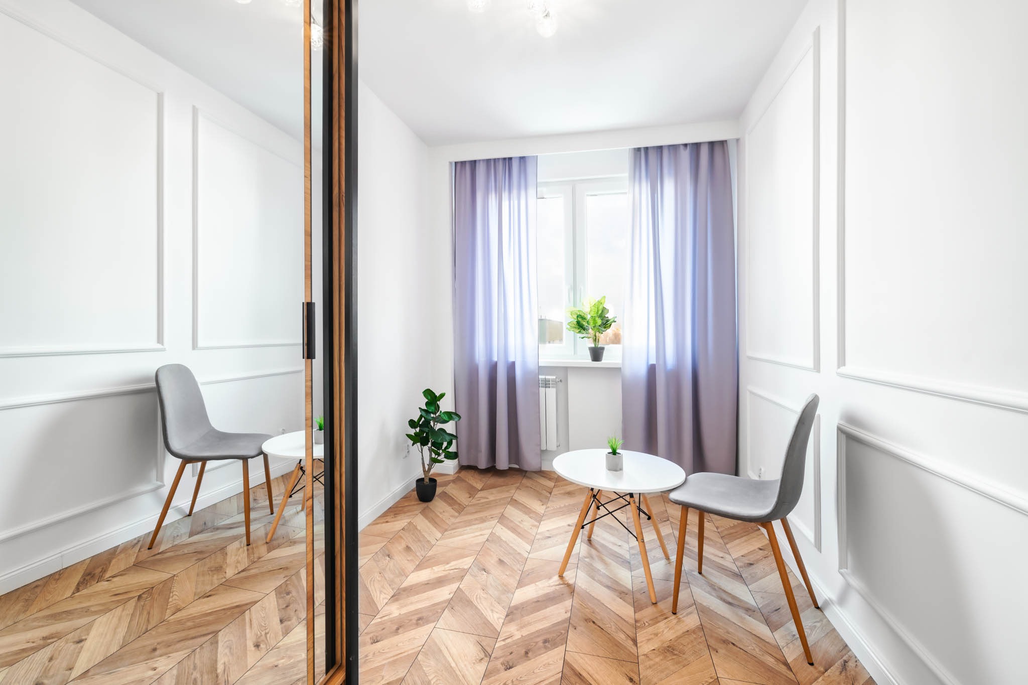 Jasne wnętrze z parkietem ułożonym w jodełkę, białe ściany z dekoracyjnymi listwami, okno z fioletowymi zasłonami, okrągły stolik i szare krzesła, lustro z drewnianą ramą.