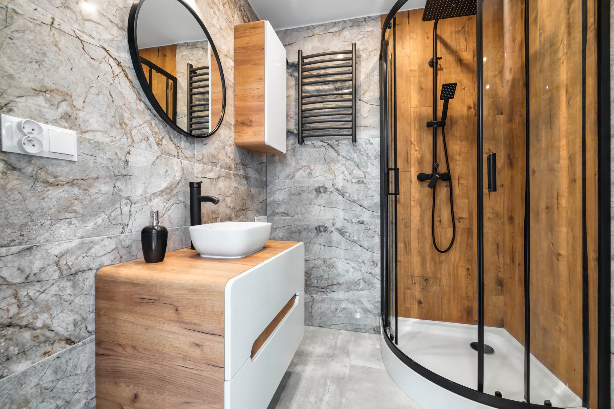 Nowoczesna łazienka z marmurowymi płytkami, drewnianą szafką pod umywalką z białą umywalką nablatową, czarną armaturą, kabiną prysznicową z drewnopodobnymi panelami oraz grzejnikiem drabinkowym.