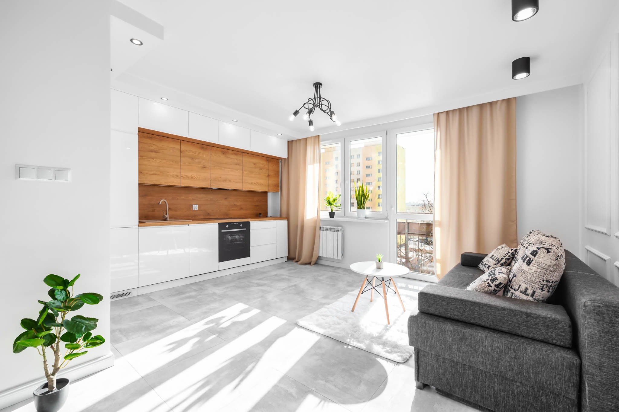 Jasne wnętrze salonu z aneksem kuchennym, białe szafki z drewnianymi frontami, szara sofa z poduszkami w gazety, szare płytki podłogowe, widok z balkonu.