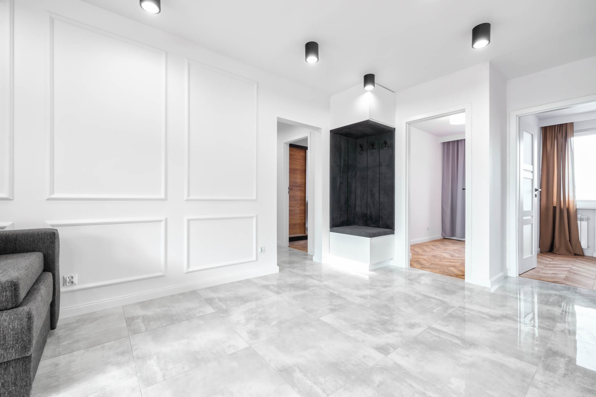 Jasne, przestronne wnętrze mieszkania z szarymi płytkami podłogowymi, białymi ścianami z dekoracyjnymi panelami, wbudowaną garderobą z podświetleniem LED i widokiem na inne pomieszczenia...