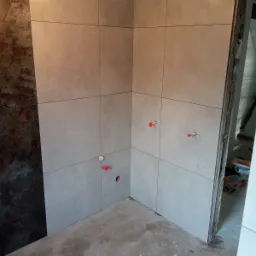 Fragment łazienki w trakcie prac: szare, kwadratowe płytki ścienne ułożone w regularną siatkę, widoczne przyłącza wodne z czerwonymi elementami, surowa podłoga i fragment ściany z surowym...