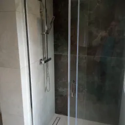 Nowoczesna kabina prysznicowa z przesuwnymi drzwiami, zamontowana armatura natryskowa z deszczownicą, odpływ liniowy w posadzce, ściany wyłożone płytkami w odcieniach szarości i ciemnej zieleni.