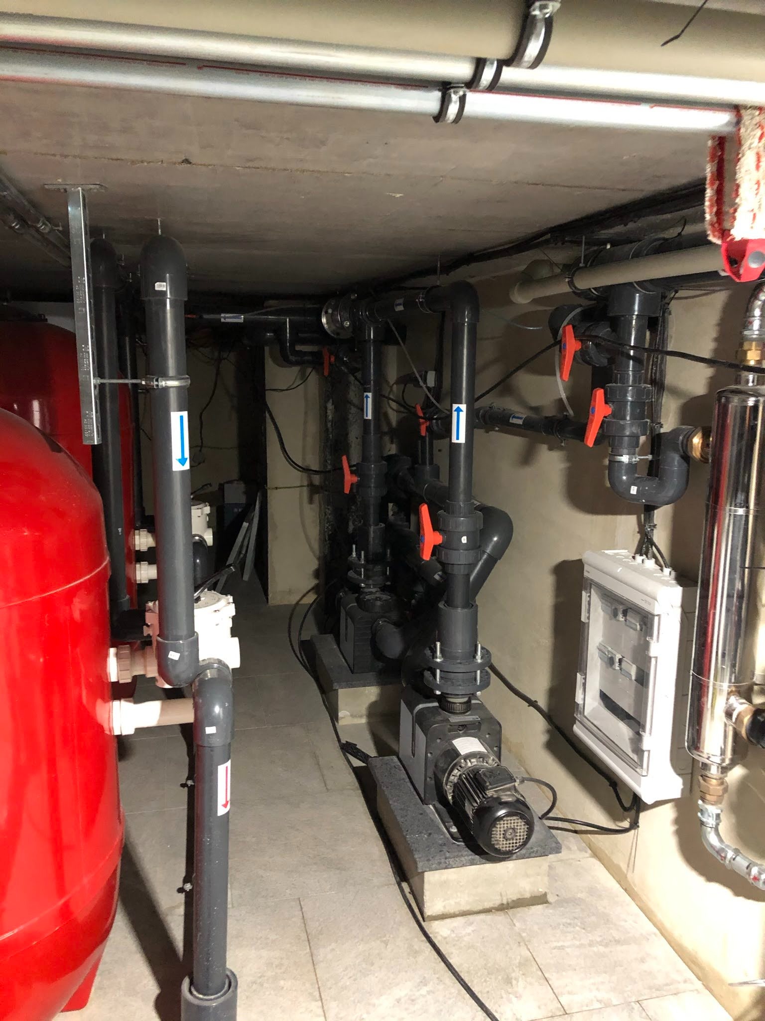 Pomieszczenie techniczne z instalacją filtracyjną i pompami do basenu, widoczne liczne rury, zawory i zbiornik ciśnieniowy w kolorze czerwonym.