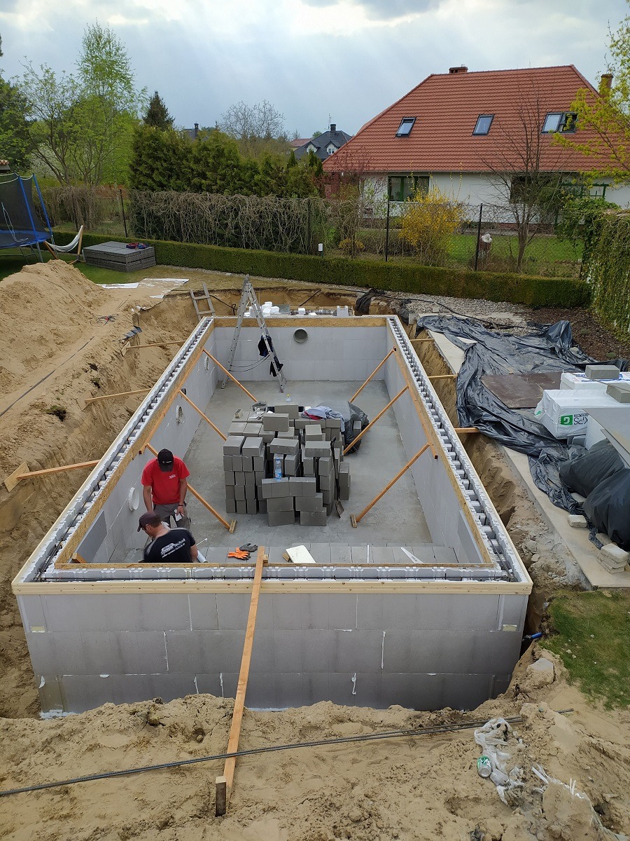 Budowa betonowego basenu w ziemi: widok z góry na wykop z ułożonymi blokami betonowymi, dwóch robotników wewnątrz, przygotowane materiały budowlane i narzędzia, dom w tle.
