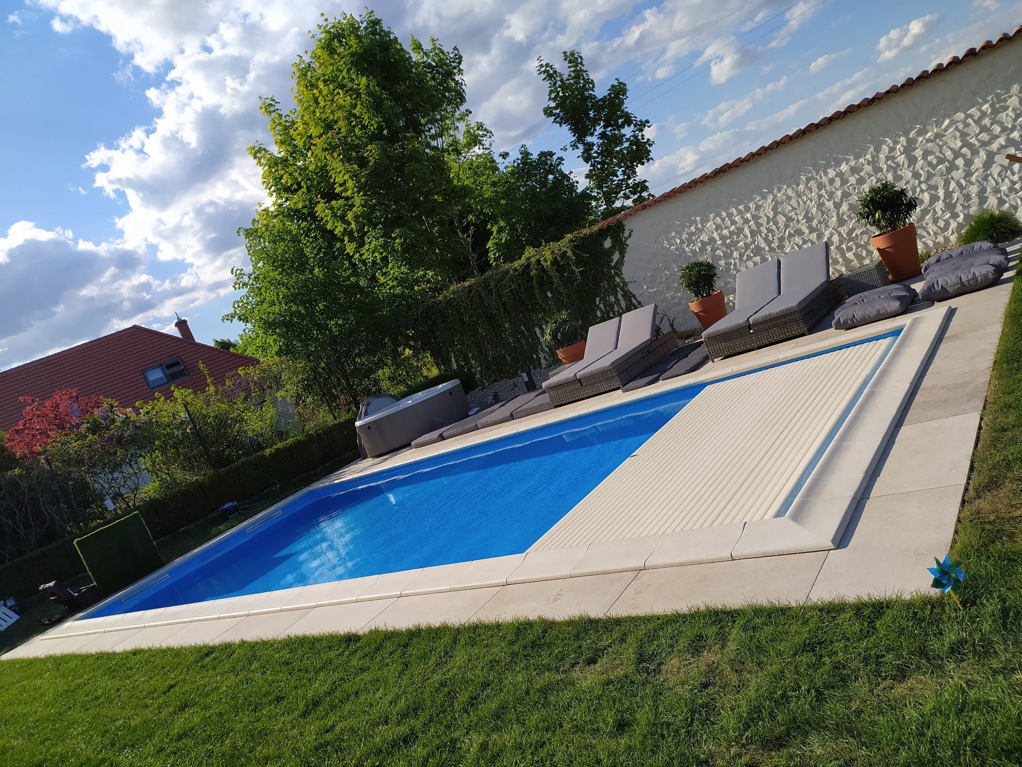 Niebieski basen z częściowo zwiniętą roletą, obok leżaki, jacuzzi i dekoracyjny wiatraczek na trawniku, w tle ogród i biały mur.