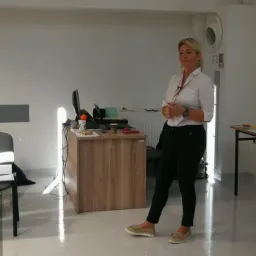 Kobieta w białej koszuli prezentuje coś w jasnej sali szkoleniowej, obok biurko z monitorem, na tablicy widoczne notatki.